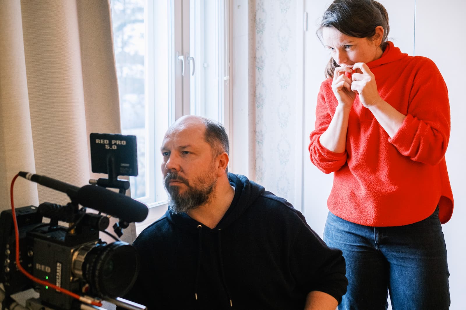 A man operates a professional video camera while a woman in a red sweater stands beside him, appearing focused and thoughtful near a window.