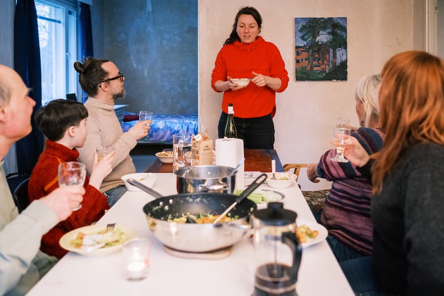 Six people sit around a dining table with food and drinks, as a woman in a red sweater stands and speaks, holding a plate.