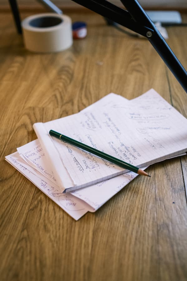 A pencil rests on handwritten notes scattered on a wooden floor, with a roll of tape and part of a metal structure visible in the background.