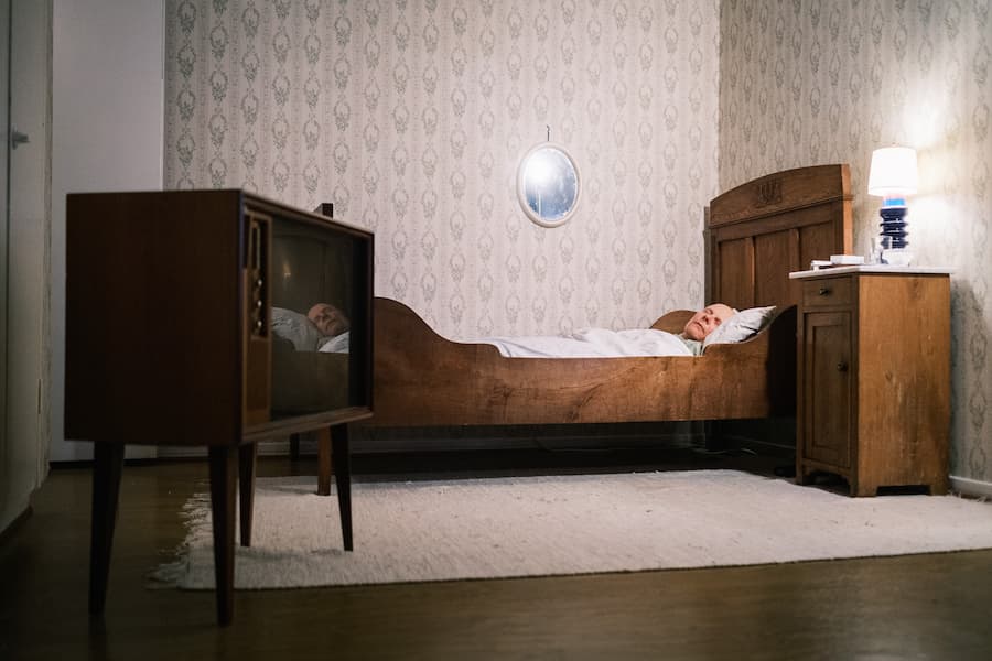 A person lies in bed in a vintage-style bedroom with patterned wallpaper, an old television, wooden furniture, and a small round mirror on the wall.