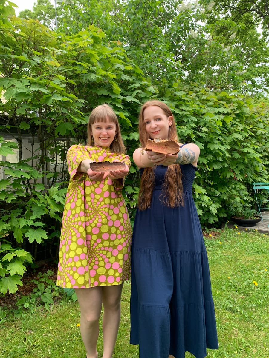 Pihla Kuulas and Kanerva Kuitu stand on grass in front of leafy bushes, smiling and holding large brown mushrooms toward the camera. One wears a patterned dress, the other a navy dress.