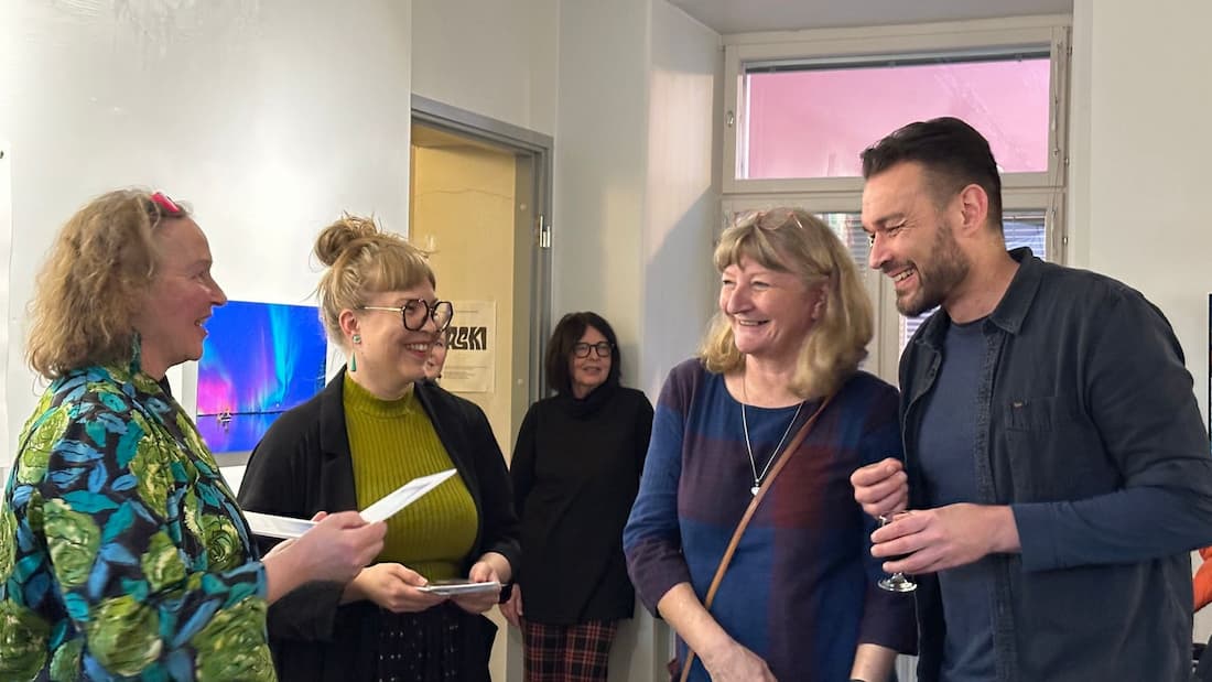 Four adults stand and talk in a brightly lit room, smiling and holding papers and a drink, while another person stands in the background near an open door.