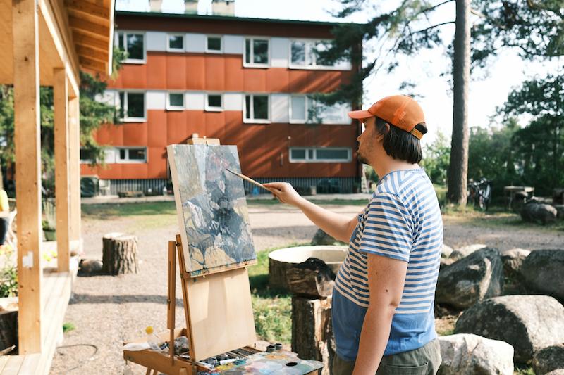 A person wearing a striped shirt and orange cap paints on an easel outdoors, with a red building and trees in the background.