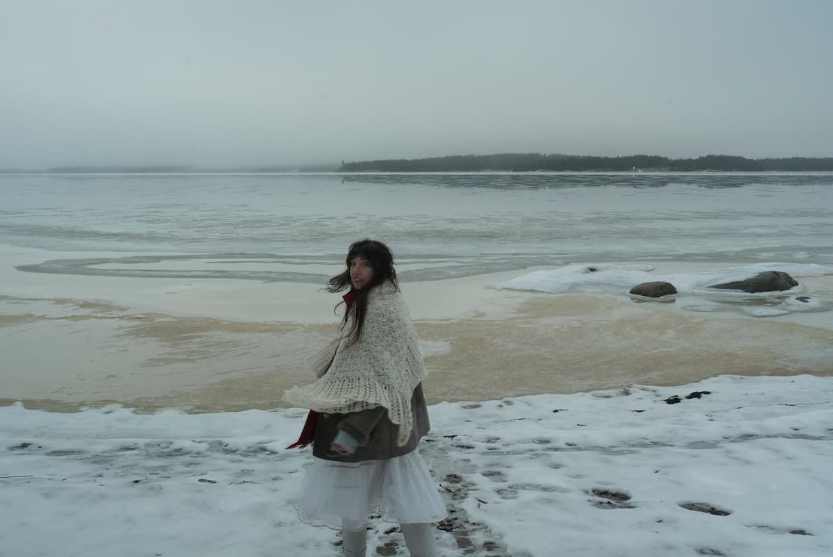 A person wearing winter clothing stands on a snowy lakeshore, looking back, with a partially frozen lake and a cloudy sky in the background.