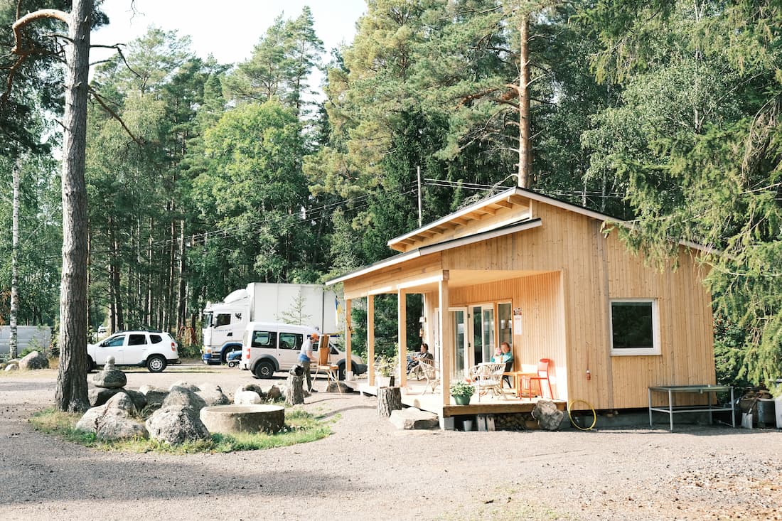 A small wooden cabin stands near several parked vehicles in a forested area on a sunny day.